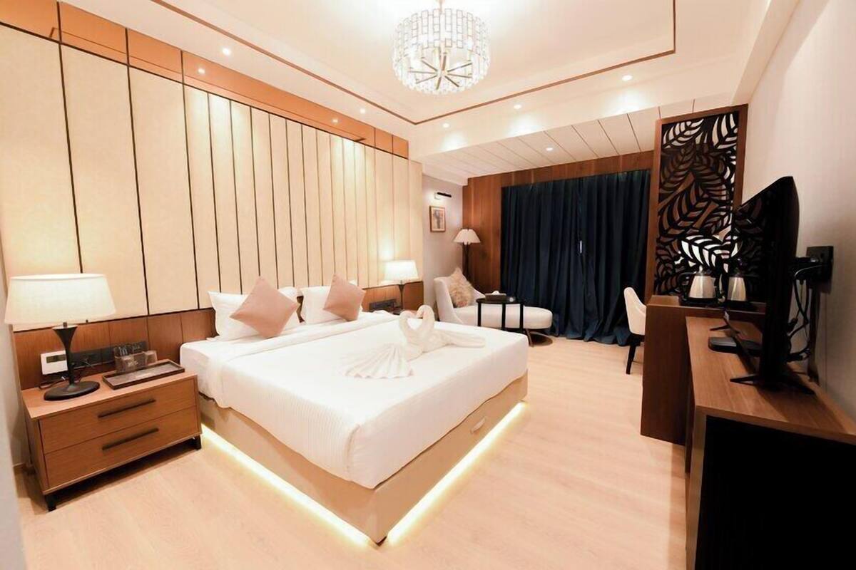 A modern hotel room with a large bed, bedside tables, a desk, a chair, a desk lamp, a decorative wall panel, and a chandelier.