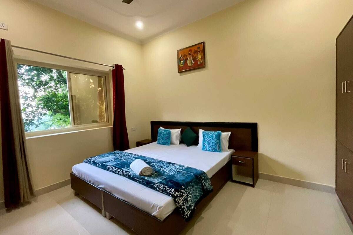 A bedroom with a bed, a window with curtains, a ceiling fan, and a painting on the wall.