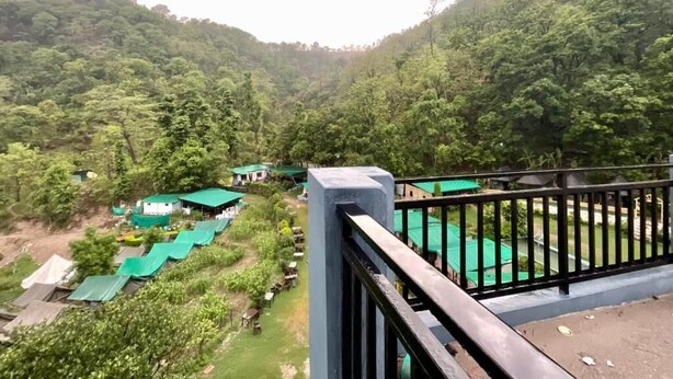 A view of a lush green valley with a building with a green roof and a black railing in the foreground. - Hotel The Rudram Aura  (Rishikesh)