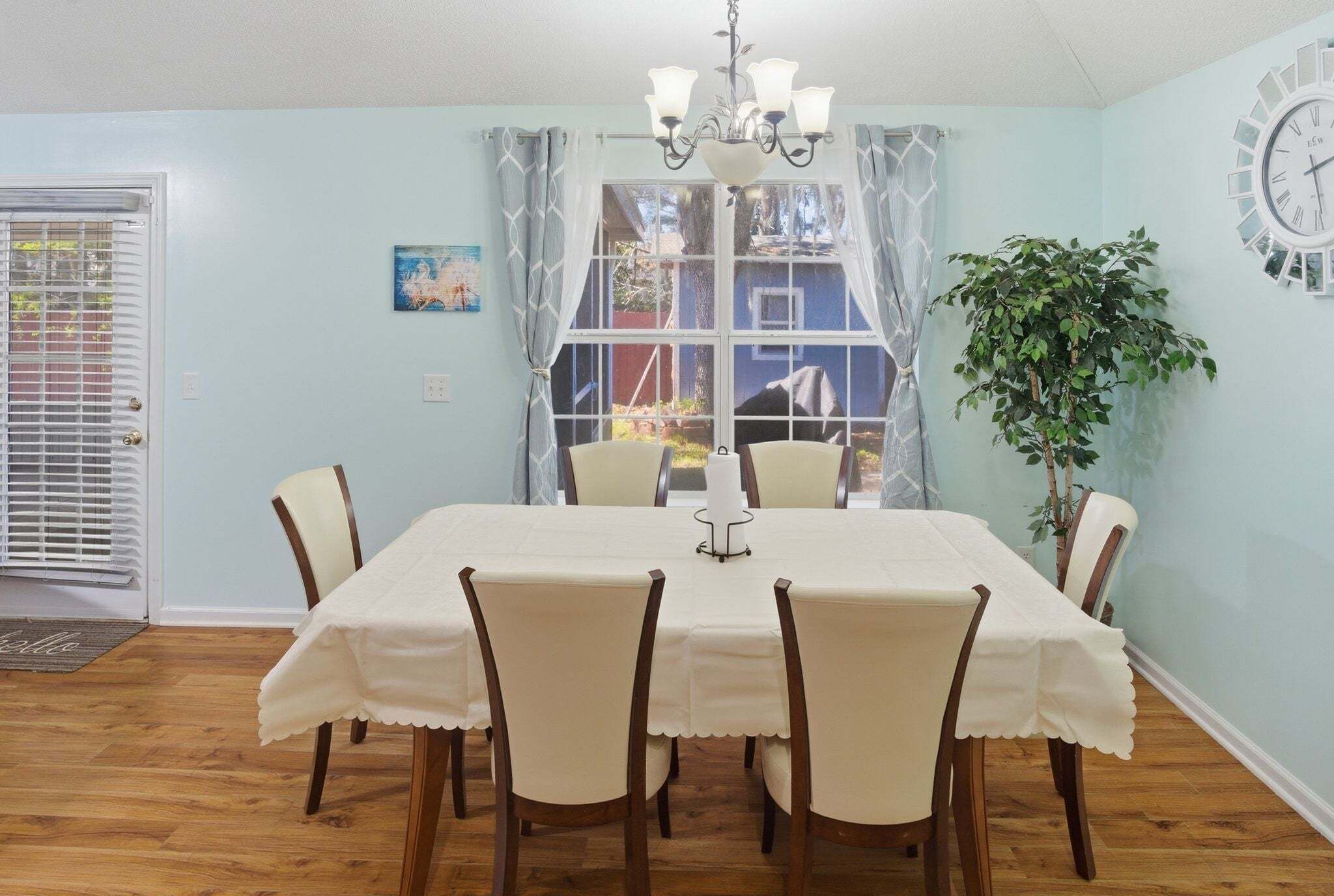 Un comedor con una mesa dispuesta para seis personas, una araña y un amplio ventanal con cortinas.