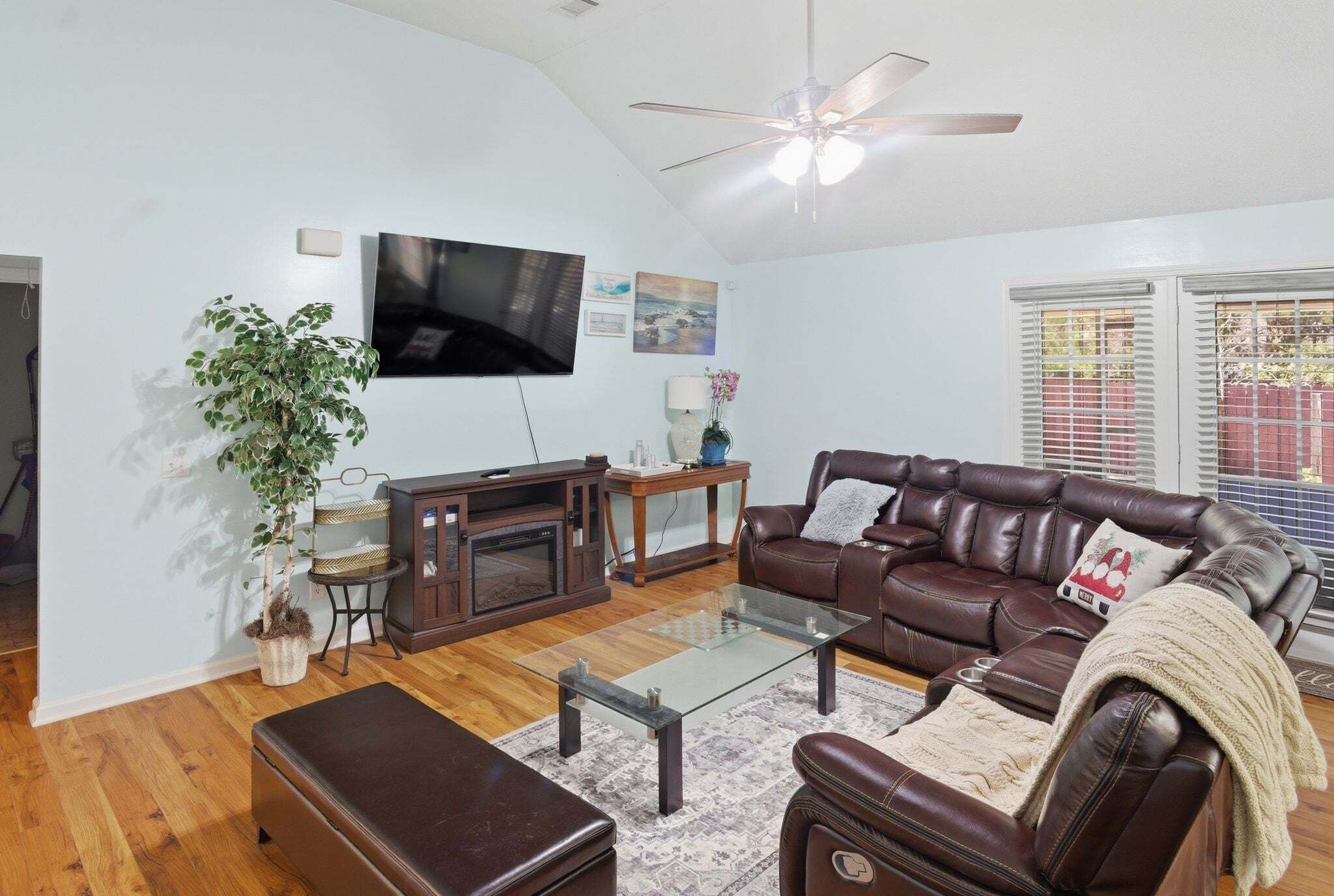 A living room with a brown leather sofa, a glass coffee table, a fireplace, a ceiling fan, and a television mounted on the wall.