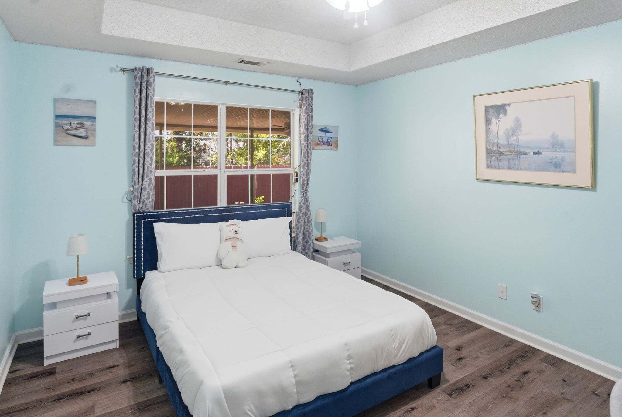 Ein Schlafzimmer mit einem Bett, einem Fenster mit Vorhängen, einem Nachttisch und einem an der Wand hängenden Bild.
