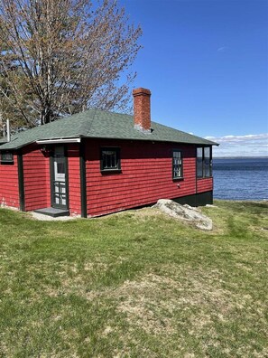 A red wooden house with a chimney, a door, and windows, located near a body of water. - Trainor Camp Limit 4 2 Bedroom Cabin (Sebago)