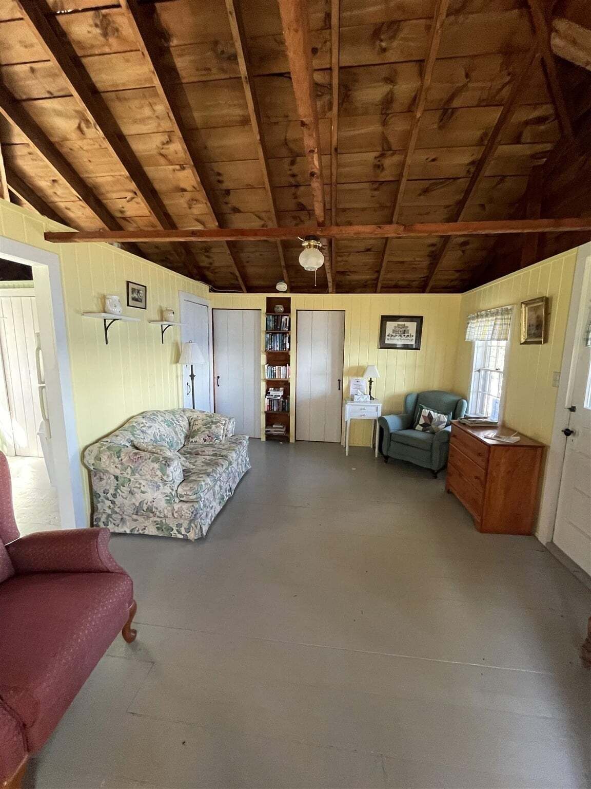 Una habitación con techo de madera, un sofá floral, un sillón rojo, un sillón azul, un aparador de madera y una estantería.