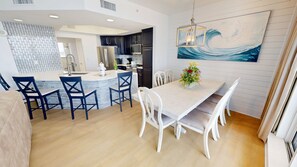 A modern kitchen with dark cabinetry, a central island, and a dining area with white chairs and a table. - Silver Beach 0704e 3 Bedroom Condo (Destin)