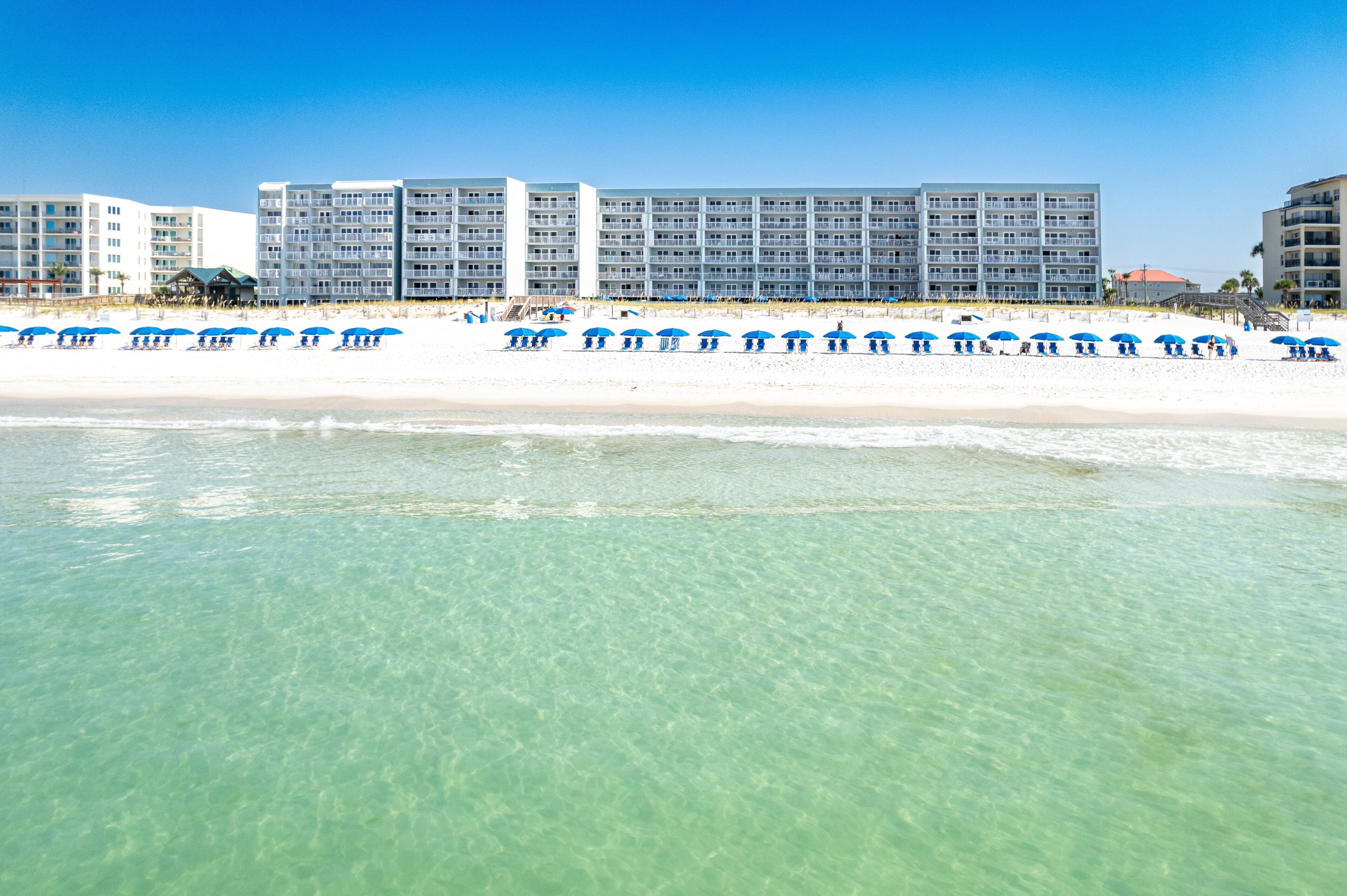 Ein Hotel direkt am Strand mit mehreren Etagen, einem Sandstrand und blauen Sonnenschirmen.