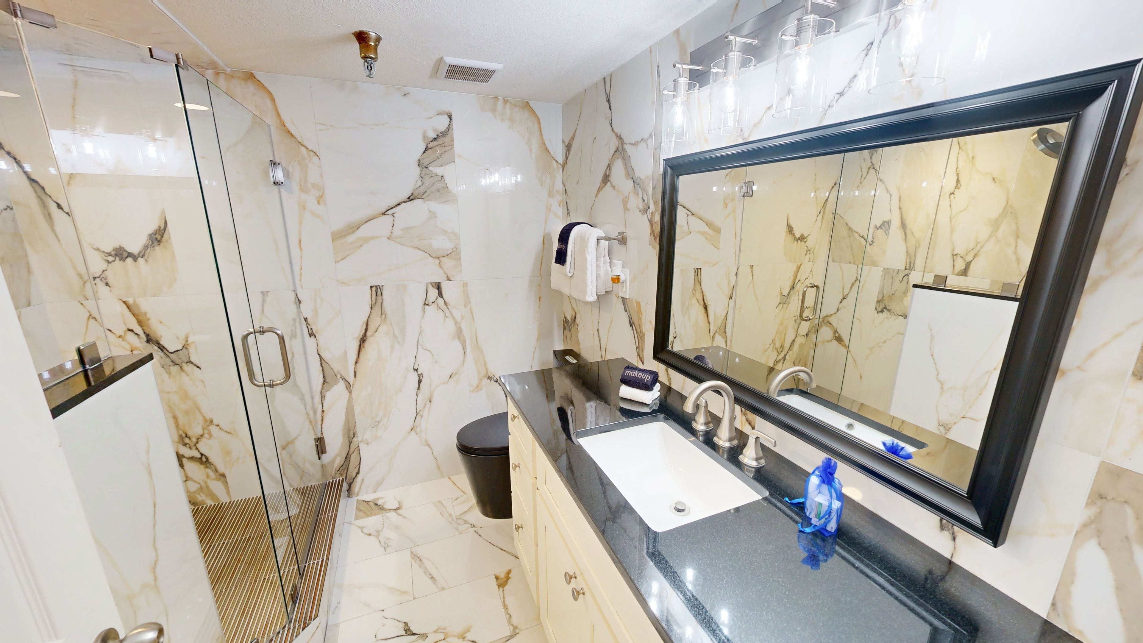 A bathroom with a marble wall, a large mirror, a sink, and a glass shower enclosure.