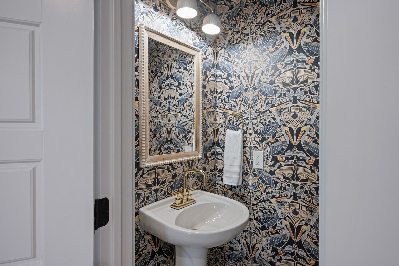 A bathroom with a patterned wallpaper, a white sink with a gold faucet, and a mirror with a gold frame.