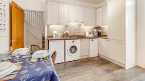 A compact kitchen with white cabinets, a washing machine, and a dining area with a table set for two. - Helens Place - North East Escapes (Whitley Bay)