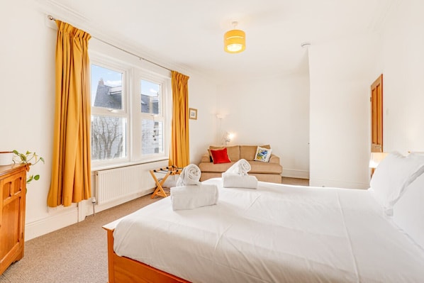 A hotel room with a bed, a sofa, a radiator, a window with curtains, and a hanging light fixture.