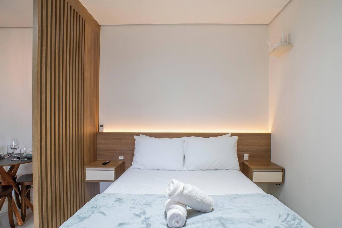 A hotel room with a bed, two bedside tables, a nightstand, a wooden partition, and a small table with glasses.