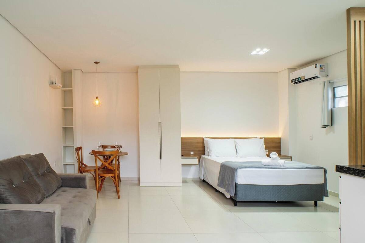 A hotel room with a bed, a sofa, a dining table with chairs, a wardrobe, and a TV mounted on the wall.