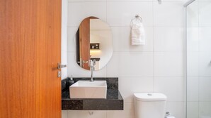 A bathroom with a wooden door, a round mirror, a white sink, a toilet, and a towel rack. - Full Studios in Rio Tavares - ALA (Florianópolis)