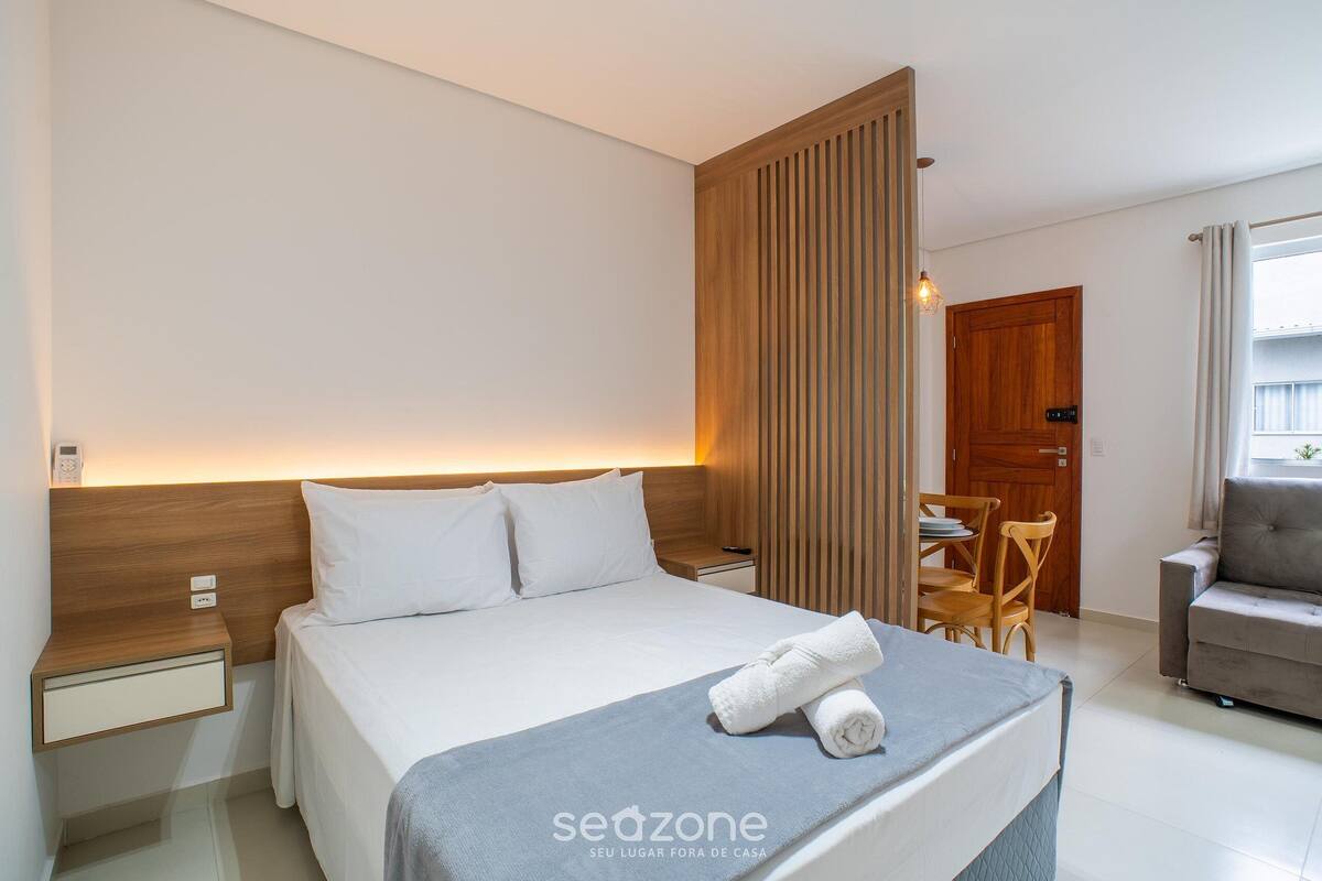 A modern hotel room with a neatly made bed, a wooden headboard, a bedside table, a chair, and a sofa.