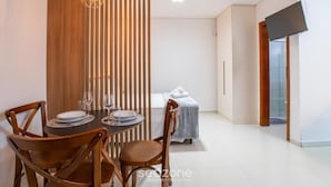 A modern room with a dining table set for two, a bed with a blue cover, and a TV mounted on the wall. - Full Studios in Rio Tavares - ALA (Florianópolis)