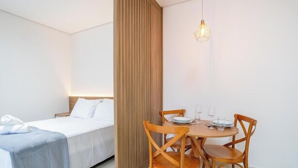 A hotel room with a bed, a wooden partition, a dining table with chairs, and a hanging light fixture. - Full Studios in Rio Tavares - ALA (Florianópolis)