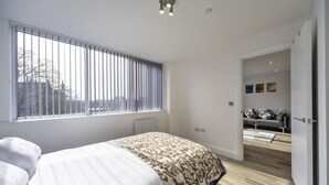 Une chambre avec une grande fenêtre, un lit recouvert d’une couvre-lit à motifs, un radiateur et une vue sur une autre pièce où se trouve un canapé.