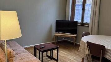 A living room with a sofa, a TV on a stand, a small table, and a dining table with chairs.
