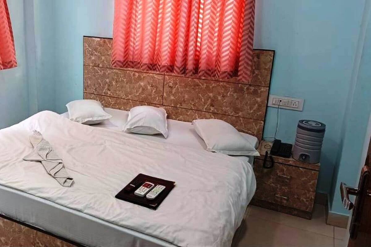 A hotel room with a bed, pillows, a bedside table with a water dispenser, and a power outlet.