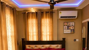 A bedroom with a bed, a ceiling fan, an air conditioning unit, and a bedside table. - Kwae Delux Hotel (Mile 11)