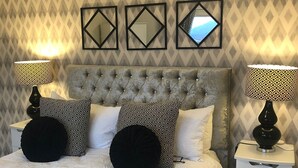 Un dormitorio con cabecera capitonada, almohadas estampadas, una mesita de noche con lámpara y un papel tapiz con diseño de diamantes.