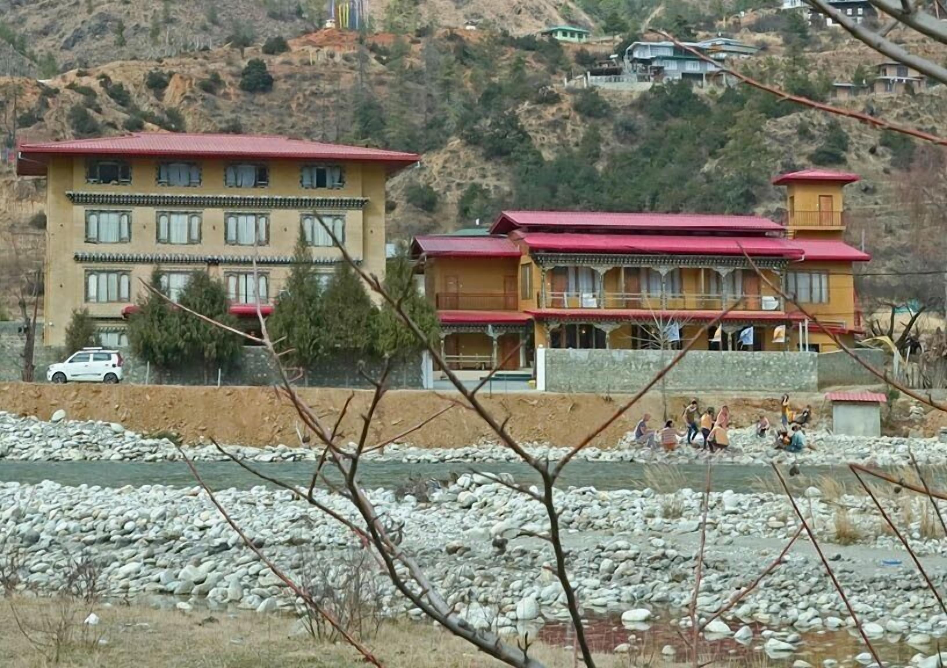 A building with multiple floors and a red roof, situated near a rocky riverbank.
