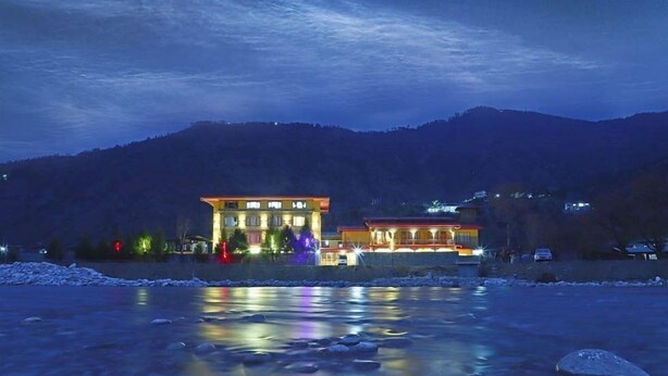 A hotel with a large building and a smaller structure across a river, surrounded by mountains. - River Front Resort (Paro)