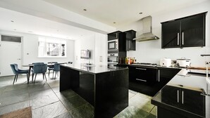 A modern kitchen with black cabinetry, a central island, and a dining area with blue chairs. - 5 Beds House - 5 Mins to Victoria Station (London)