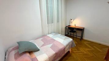 En säng för en person med ett mönstrat överkast, ett skrivbord med en stol, en lampa och ett fönster med tunna, genomskinliga gardiner.