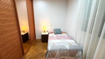 A bedroom with a bed, two bedside tables with lamps, a wooden wardrobe, and a window with sheer curtains.