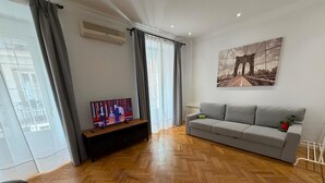 Una sala de estar con un sofá, un televisor sobre un mueble bajo, un cuadro en la pared y una ventana con cortinas.