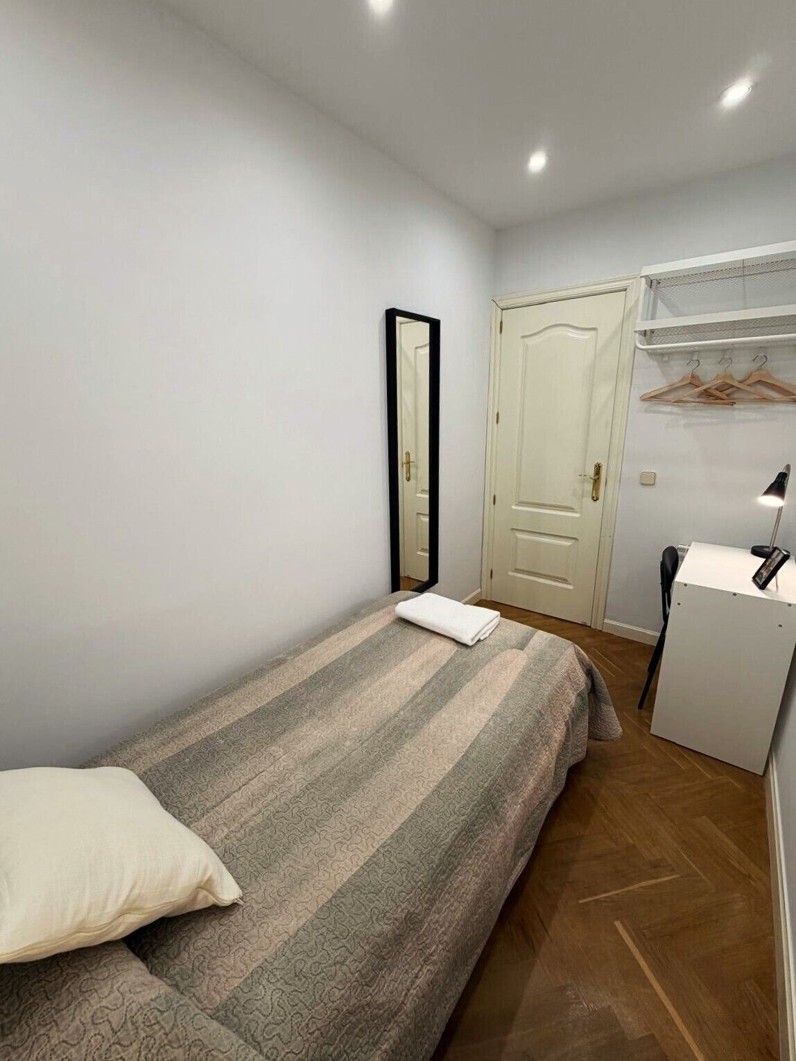 Een kleine, verzorgde slaapkamer met een bed, een spiegel, een deur en een bureau met een stoel.