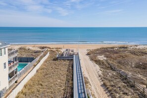 Strandudsigt med en bygning, en swimmingpool og en træbro, der fører til stranden.