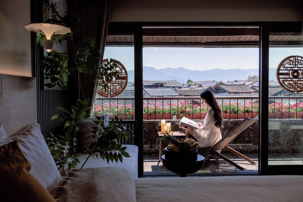 A room with a large window, a chair, a table with fruits, and a person reading a book.