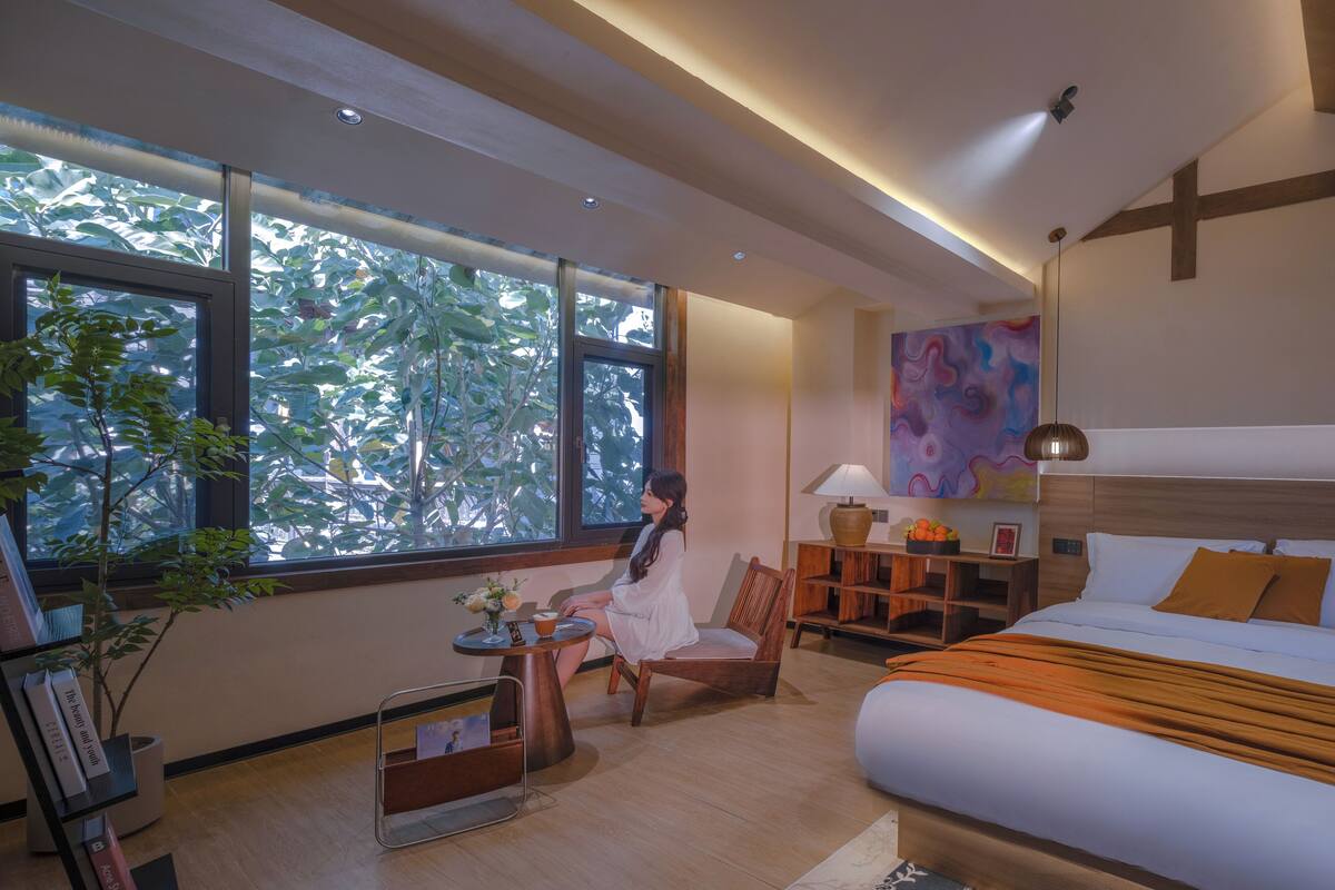 A hotel room with a large window, a bed, a chair, a small table with a lamp, and a bookshelf.