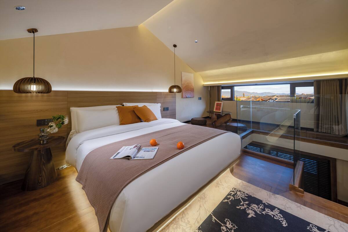 A modern hotel room with a large bed, a wooden headboard, a bedside table with a vase, and a view of the city through a balcony.