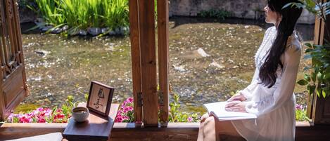Eine Person sitzt am Fenster mit Blick auf einen Teich, an einem Tisch stehen eine Tasse, eine Schüssel und ein gerahmtes Bild.