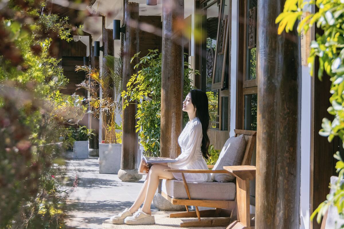 A person sitting on a wooden bench in a covered outdoor area with potted plants and a pathway.