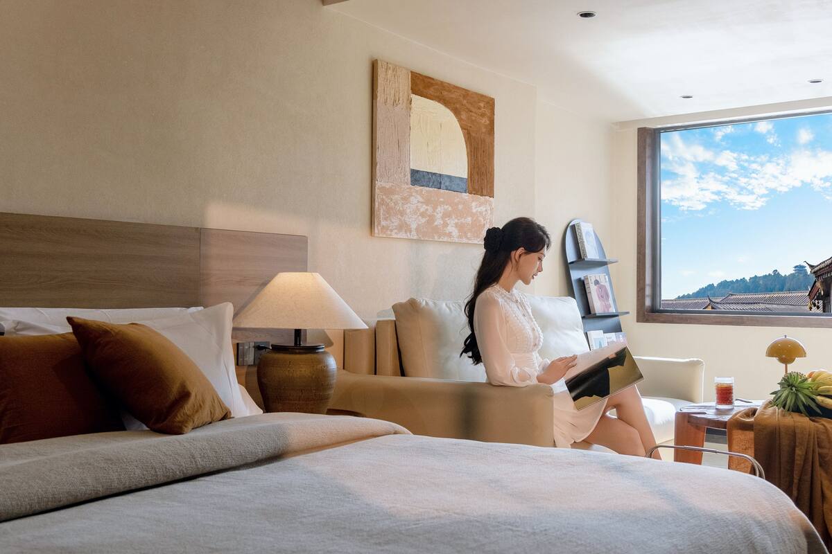 A hotel room with a bed, a sofa, a table with a lamp, a window with a view, and a painting on the wall.