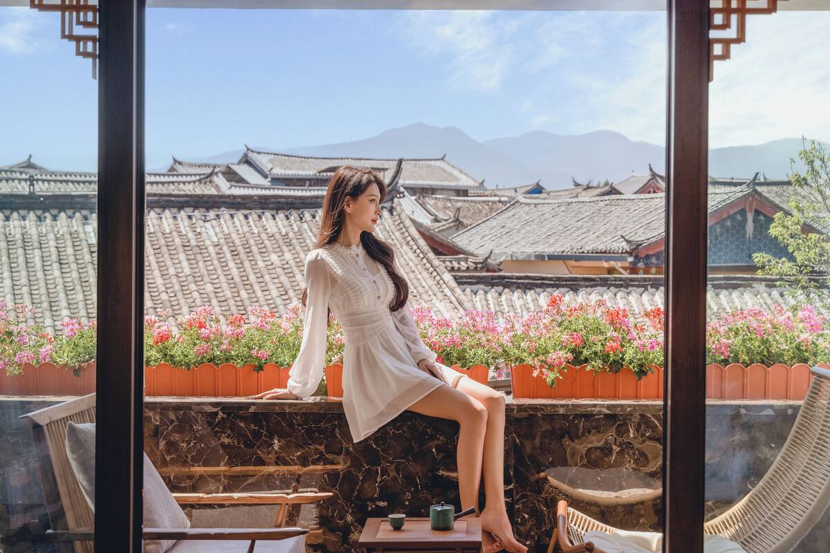 A woman sitting on a balcony with traditional architecture and potted plants in the background.