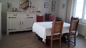 Een eetruimte met een witte tafel, houten stoelen, een wit kastje met decoratieve knoppen, een ronde spiegel en een hanglamp.