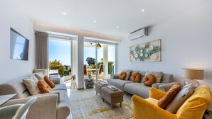 A spacious living room with a sofa set, a coffee table, and a yellow armchair. There is a balcony with outdoor furniture visible through the glass doors. - 1D Waterside with Shared Pool (Luz)