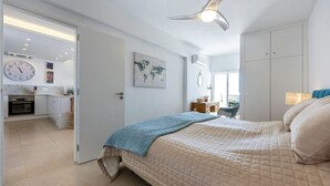 A bedroom with a bed, a ceiling fan, a world map on the wall, and a kitchen area visible through an open door. - 1D Waterside with Shared Pool (Luz)