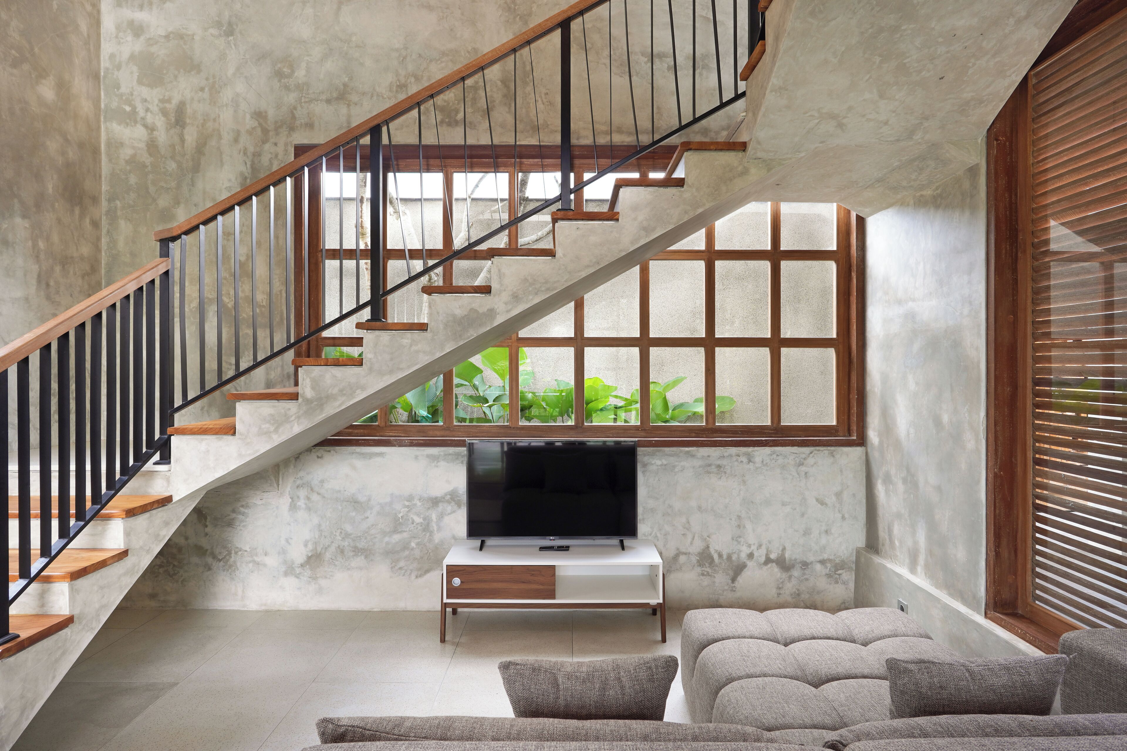 A living room with a staircase, a flat-screen TV on a white stand, a grey sofa, and a window with blinds.