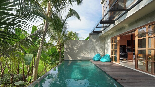 Un área de piscina moderna con cubierta de madera, cocina con paredes de vidrio y exuberante vegetación tropical.
