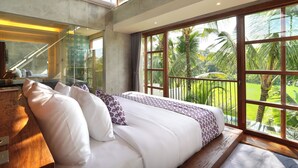 Un dormitorio con una cama grande, un escritorio de madera, un espejo y vistas a un jardín exuberante a través de amplios ventanales.