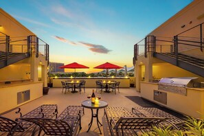 An outdoor patio with tables and chairs, red umbrellas, a grill, and a sunset view. - Amazing Location, Close 2 Seaport Village W/ Gym & Parking! (San Diego)