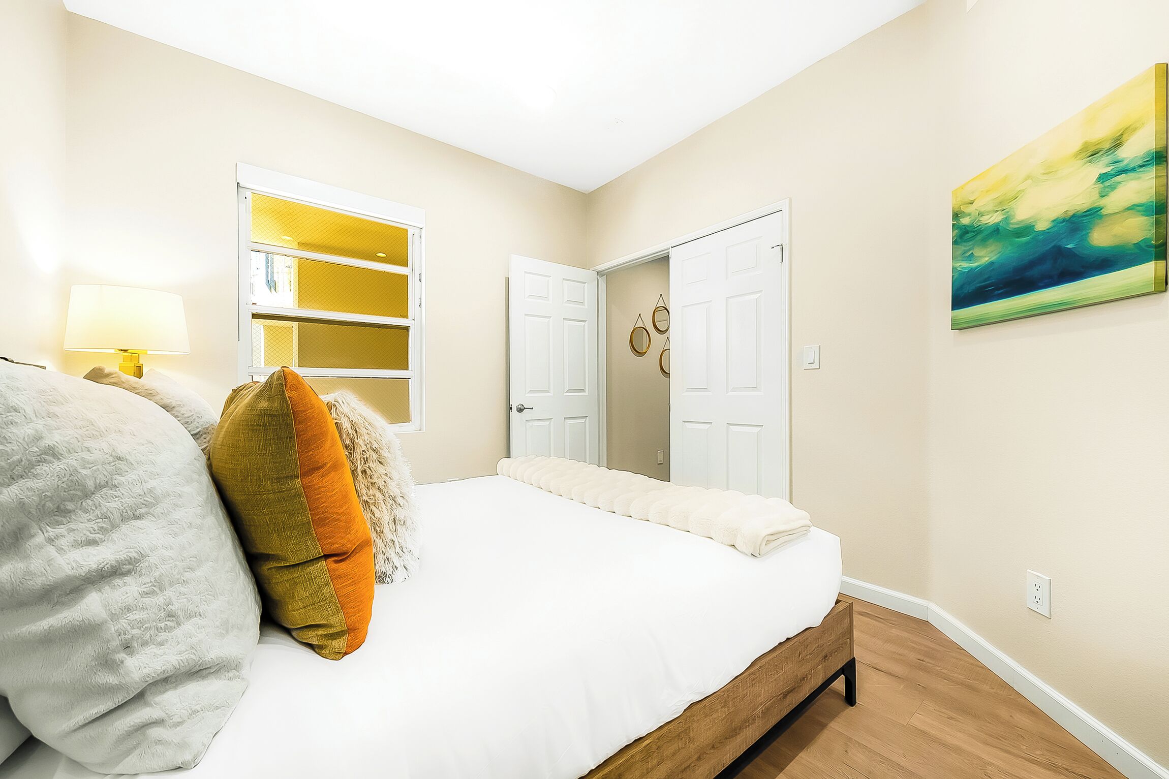 Une chambre à coucher avec un lit, une table de chevet, une lampe, une fenêtre, une porte et un tableau au mur.