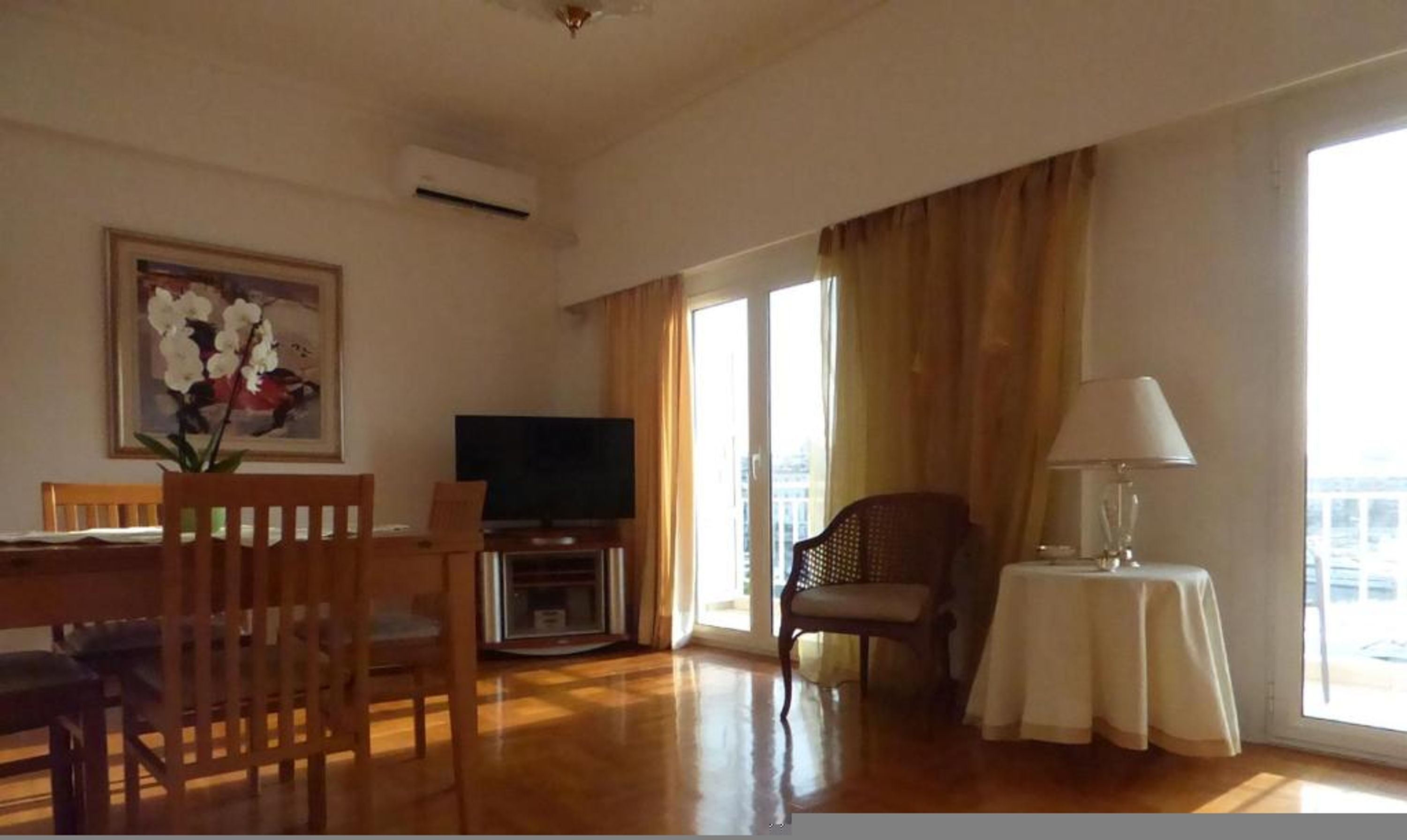 Una stanza con un tavolo da pranzo in legno, sedie, una televisione, una lampada e un'ampia finestra con tende.