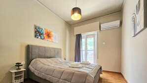 A bedroom with a bed, a nightstand, a potted plant, a window with curtains, and an air conditioning unit. - El Prestige Apartment - Athens Town (Athens)